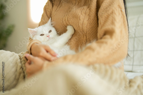 Young Asian woman in brown sweater sitting on bed and embrace her white fluffy kitten , looking at her with love emotion. Bonding between pet and owner concept.