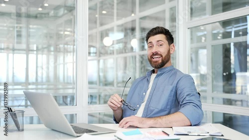 Happy businessman is satisfied with completed task on laptop while sitting at desk at workplace in a modern business office. Smiling worker successfully finished work on project and looking at camera