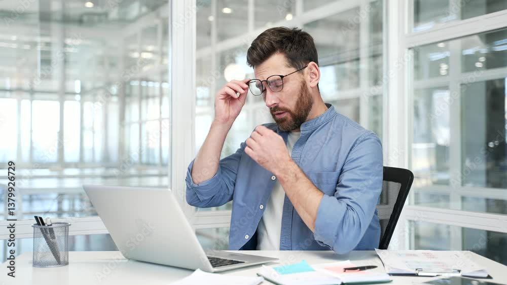 Overworked businessman suffering from eye strain while working on a laptop computer sitting at workplace in modern business office. Tired male worker takes off glasses and massages rubs bridge of nose Overworked businessman suffering from eye strain while working on a laptop computer sitting at workplace in modern business office. Tired male worker takes off glasses and massages rubs bridge of nose