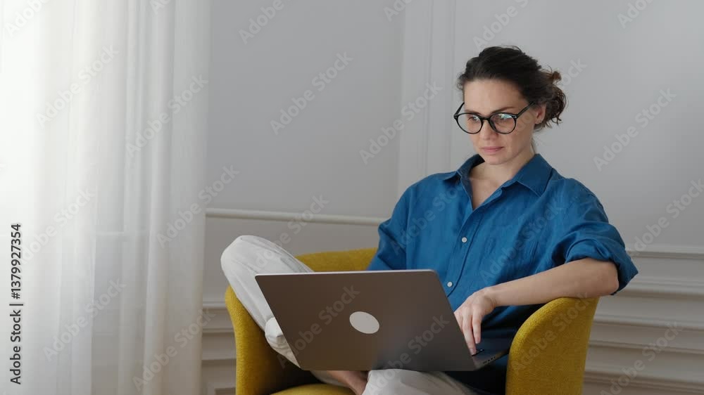 A young woman is working on a laptop while sitting in an armchair in the living room