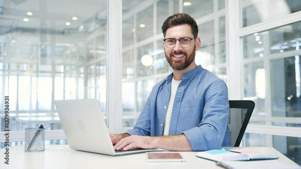 Portrait of happy businessman sitting at desk at workplace in modern business office. Confident successful smiling entrepreneur using laptop and looking at camera. Handsome worker works at workspace Portrait of happy businessman sitting at desk at workplace in modern business office. Confident successful smiling entrepreneur using laptop and looking at camera. Handsome worker works at workspace
