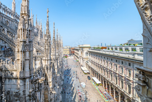 Fototapeta Naklejka Na Ścianę i Meble -  A breathtaking panorama from the rooftop of Duomo, showcasing the intricate architecture of the cathedral alongside bustling streets and nearby buildings in Milan on a clear day.