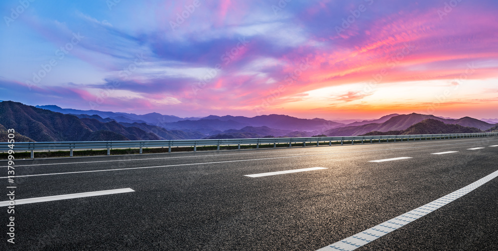 Empty asphalt highway leads towards stunning mountain range at colorful sunset