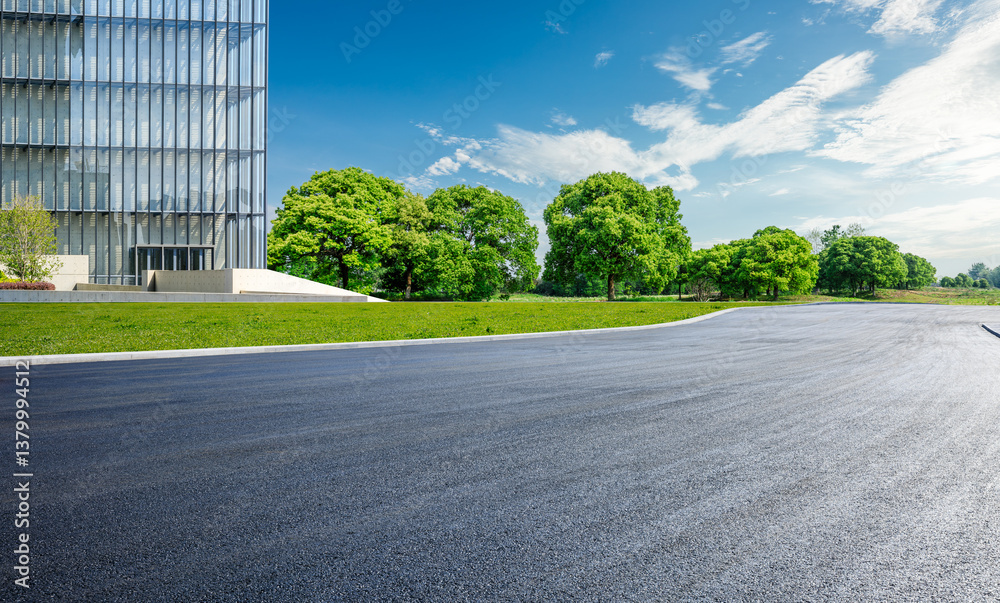 Empty asphalt road curves past a modern glass office building and green park trees under the blue sky