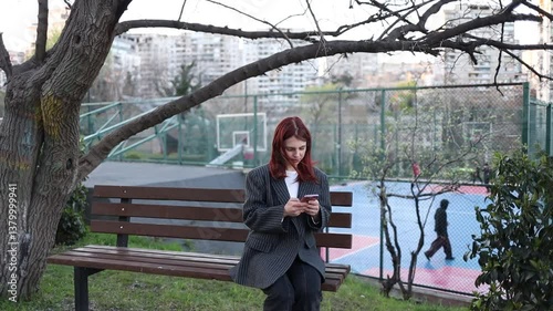 A girl sits on a bench in a park and types on her phone. A red-haired woman with a phone is relaxing on a bench in a city park. A woman works on a phone in a park. Remote work on the phone.