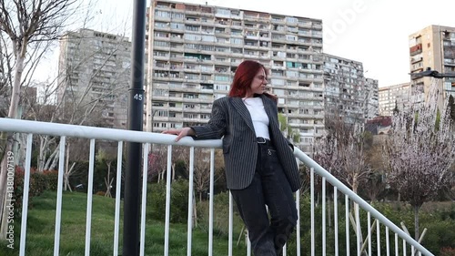 Beautiful girl against the backdrop of multi-story buildings. Red-haired girl leaning on the railing. Woman in a business suit walking in the park.
