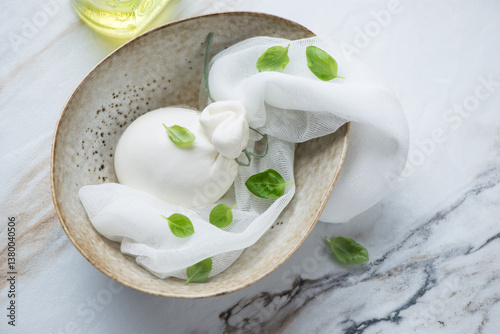 Bowl with burrata cheese in a brine and basil leaves, horizontal shot on a white stone background, high angle view