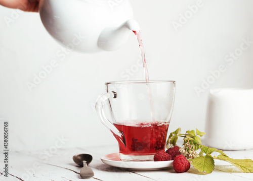 Female hand holding a teapot and pouring tea into a transparent glass cup with a raspberries. On a white plate on a wooden background.