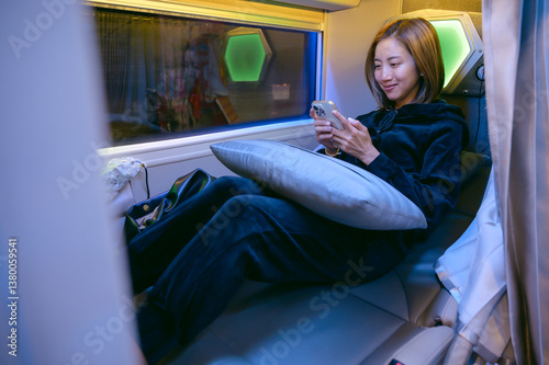 Fotografija A young woman relaxing on a sleeper bus at night while using her smartphone, enjoying solo travel comfort