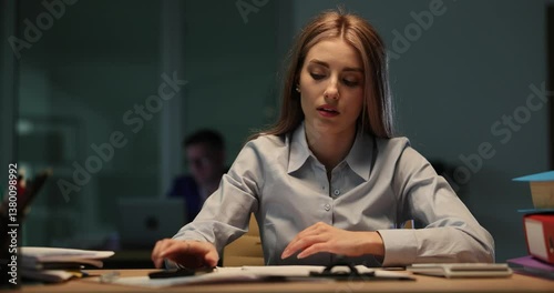 Woman with phone slumped over desk appearing tired and stressed in office setting. Emphasizing late and overtime work environment in evening