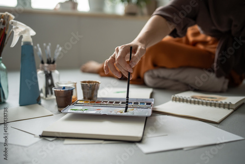 Unrecognizable woman artist hand mixing watercolors for sketch in notebook on floor in studio. Nearby sprayer and small cups to assist with artwork, tools ready for creative work, color palette idea