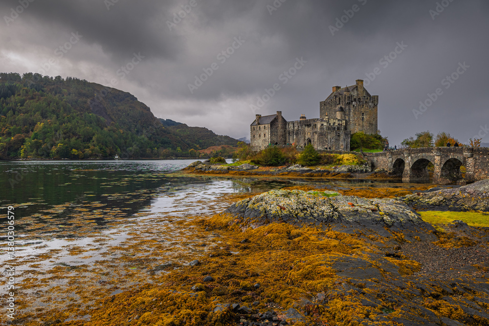 Eilean Donan Castle stands majestically amid autumn hues, its reflection shimmering in calm waters. Misty Highlands and golden trees create a scene of timeless beauty.