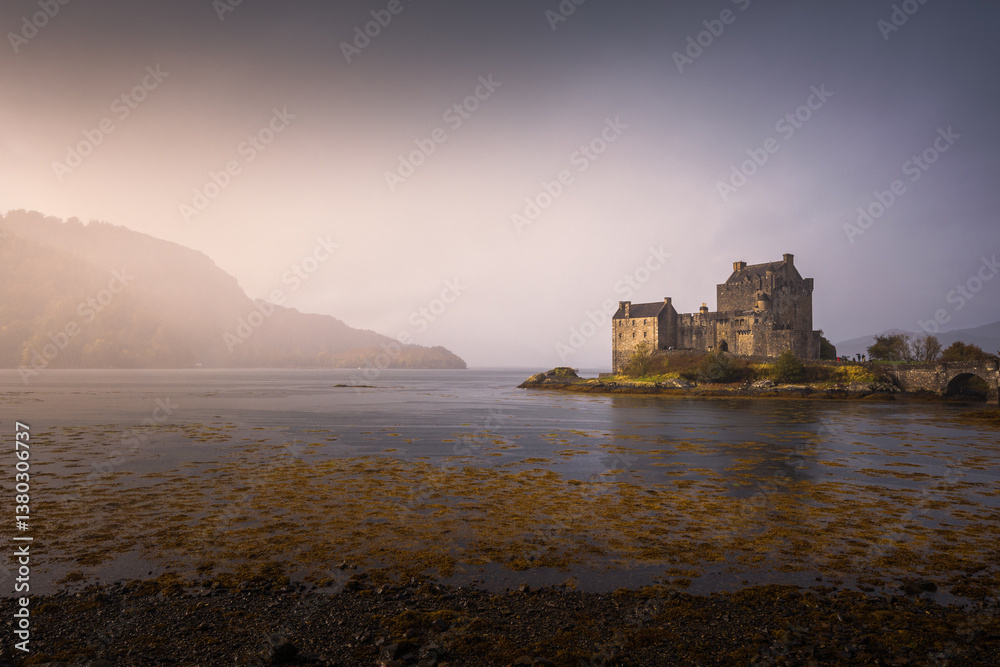Eilean Donan Castle stands majestically amid autumn hues, its reflection shimmering in calm waters. Misty Highlands and golden trees create a scene of timeless beauty.