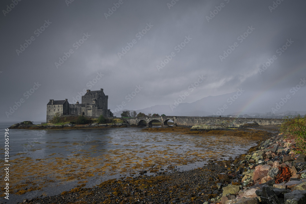 Eilean Donan Castle stands majestically amid autumn hues, its reflection shimmering in calm waters. Misty Highlands and golden trees create a scene of timeless beauty.