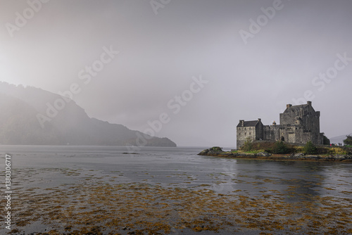 Eilean Donan Castle stands majestically amid autumn hues, its reflection shimmering in calm waters. Misty Highlands and golden trees create a scene of timeless beauty.