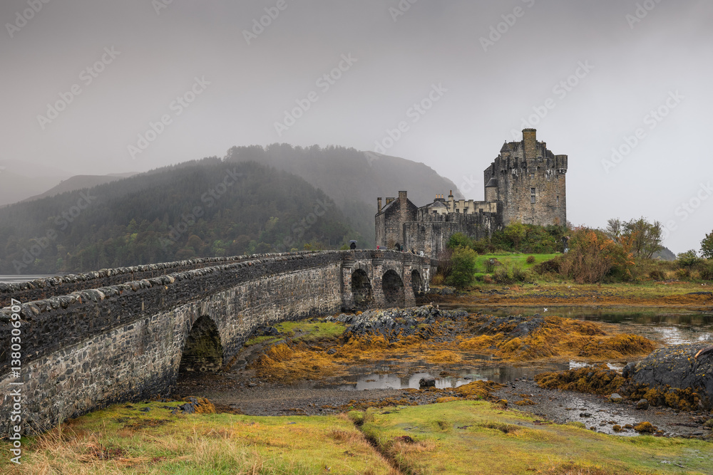Eilean Donan Castle stands majestically amid autumn hues, its reflection shimmering in calm waters. Misty Highlands and golden trees create a scene of timeless beauty.