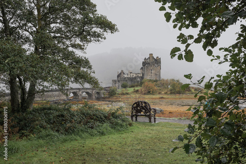 Eilean Donan Castle stands majestically amid autumn hues, its reflection shimmering in calm waters. Misty Highlands and golden trees create a scene of timeless beauty.