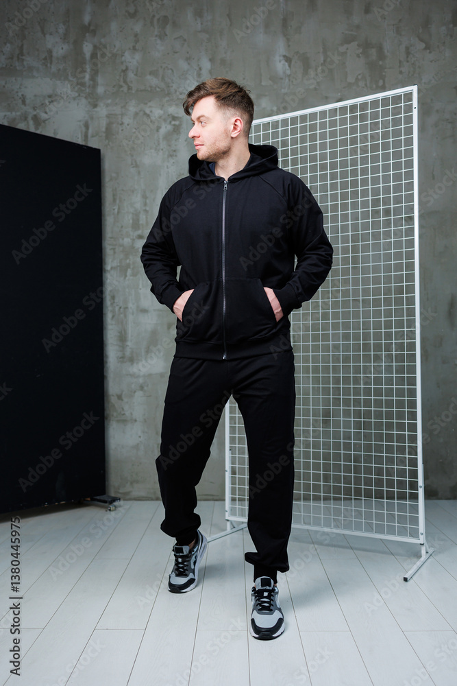 Obraz premium A Stylish Young Man Dressed in Trendy Black Sportswear Is Posing for a Fashion and Lifestyle Photography Session in a Modern Studio Setting, Emphasizing His Attitude and Contemporary Style