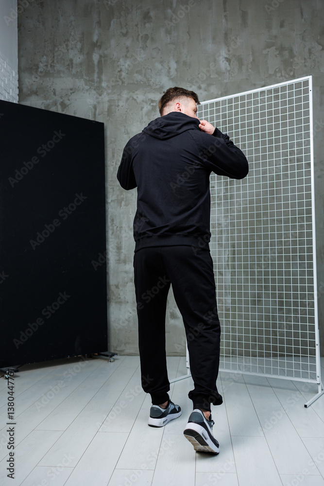 This image features a stylish young man dressed in black sportswear, confidently posing in a modern interior setting that highlights trendy streetwear fashion and makes a strong visual statement
