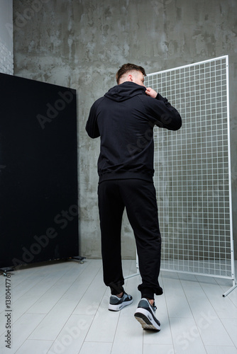 Obraz na płótnie This image features a stylish young man dressed in black sportswear, confidently posing in a modern interior setting that highlights trendy streetwear fashion and makes a strong visual statement w salonie