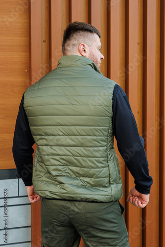 Obraz na płótnie A stylish man in an olive green puffer vest and black longsleeve shirt is seen from behind, set against a vibrant modern architectural backdrop, creating a trendy outdoor atmosphere w salonie