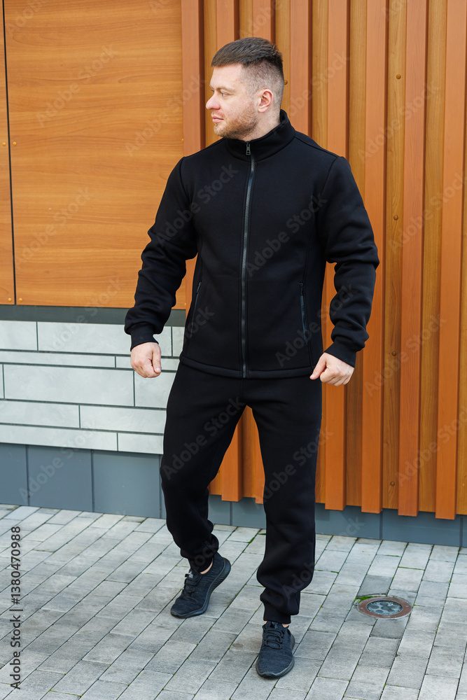 A man is confidently posing outdoors in a stylish black athletic outfit, which perfectly complements the sleek and modern architectural structure that is set nearby in the picturesque background