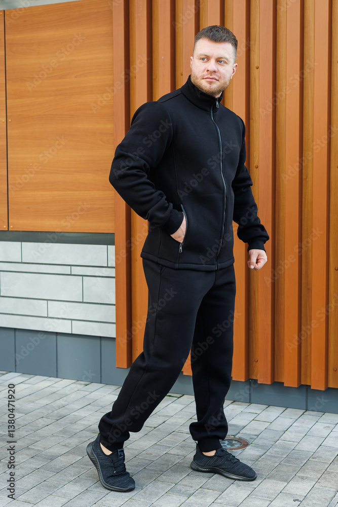 A confident young man in a stylish black outfit poses elegantly against a beautiful wooden backdrop, radiating charm and charisma, making his trendy urban style stand out in any crowd