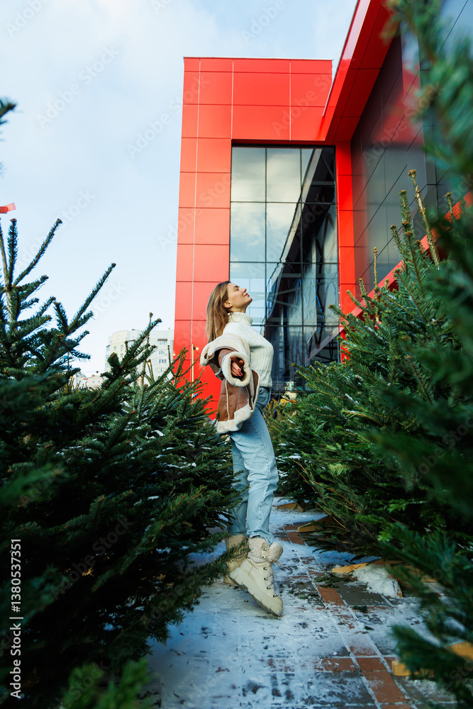 A fashionable young woman elegantly poses beside a striking red building, framed by evergreen trees, creating an inviting urban scene that captures attention and admiration from onlookers