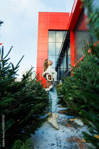 Obraz na płótnie A fashionable young woman elegantly poses beside a striking red building, framed by evergreen trees, creating an inviting urban scene that captures attention and admiration from onlookers w salonie