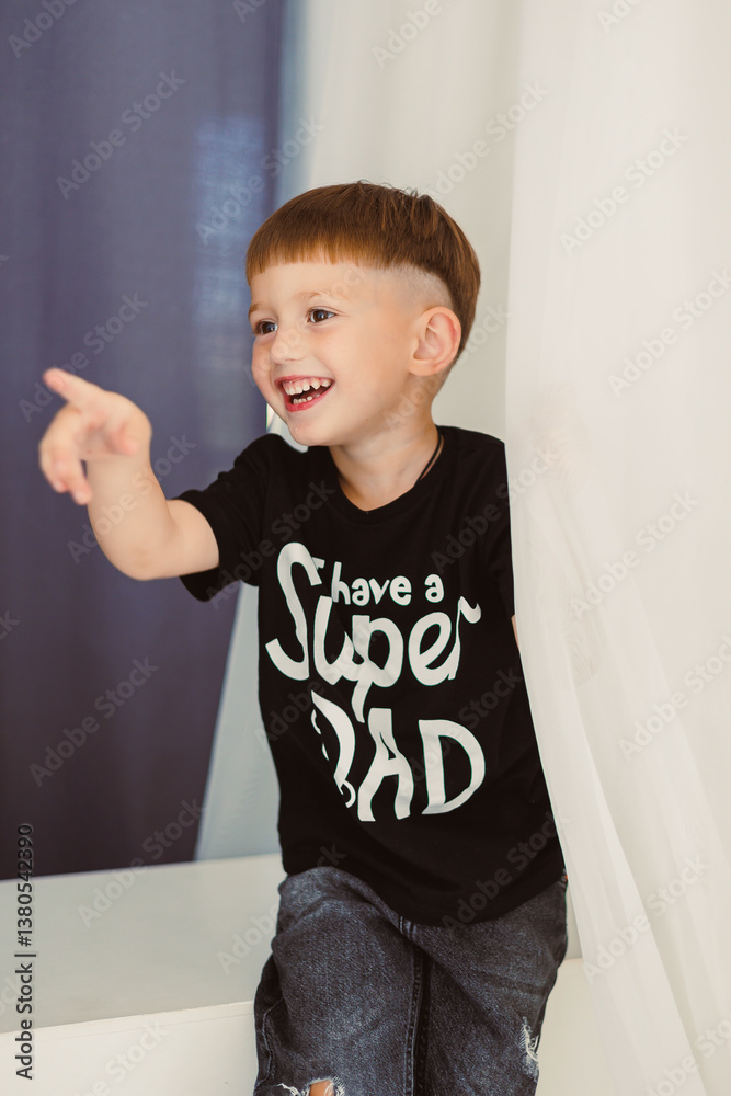 A joyful boy in a trendy Black Super Dad TShirt points excitedly, his big smile lighting up his face, showcasing his lively spirit and embodying the essence of childhood happiness and wonder