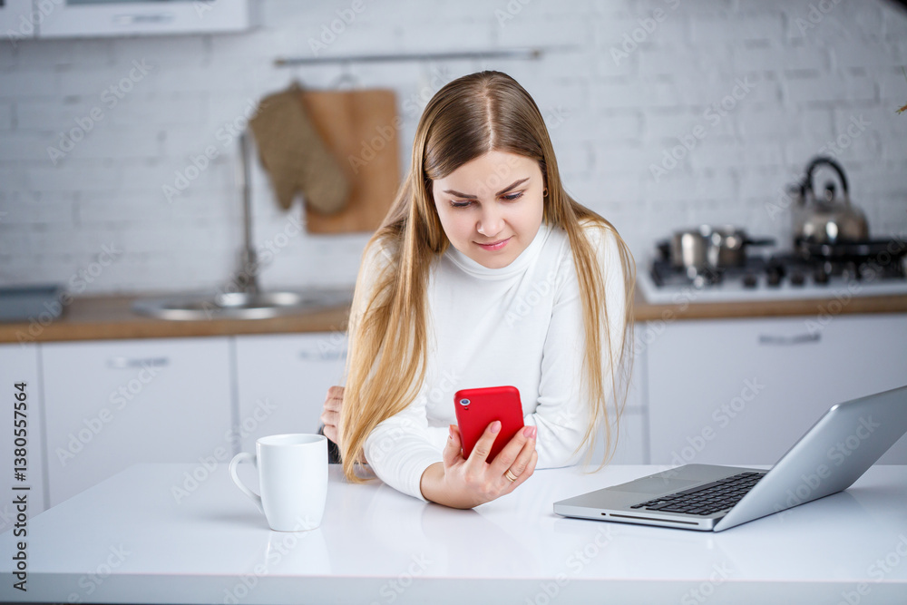 A young woman engages with her smartphone in a modern kitchen, accompanied by a laptop and coffee, creating a cozy atmosphere of relaxation and focus in a contemporary domestic lifestyle
