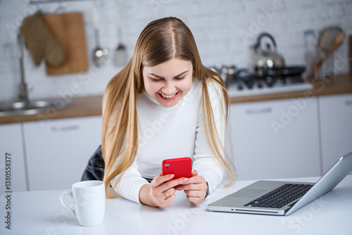 A cheerful young woman smiles as she texts friends in her modern kitchen with contemporary furniture, a laptop on the counter, and a steaming cup of coffee, reflecting her relaxed, creative lifestyle