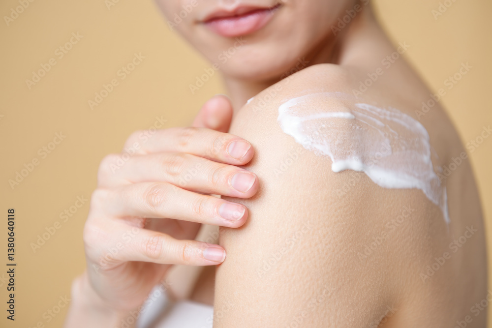Fototapeta premium Young caucasian female applying moisturizing cream to shoulder on beige background