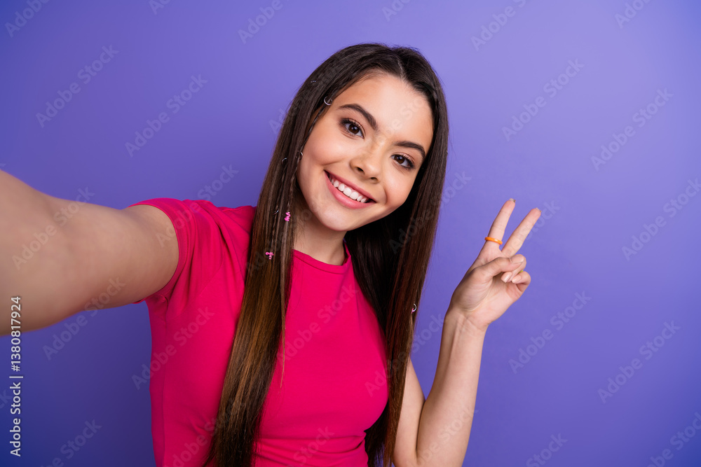 Fototapeta premium A cheerful young woman in a pink t-shirt taking a selfie and making a peace gesture against a vibrant purple background