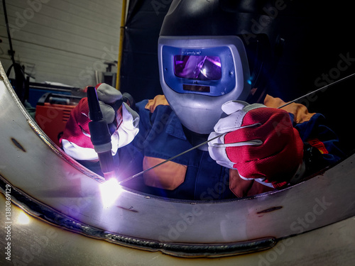 professional welder working on a metal structure using TIG (Tungsten Inert Gas) welding. The welder is wearing a protective suit, a darkened visor helmet, and red-white gloves while holding a welding 