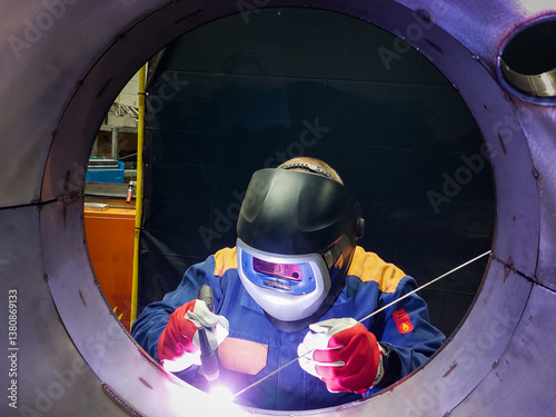professional welder working on a metal structure using TIG (Tungsten Inert Gas) welding. The welder is wearing a protective suit, a darkened visor helmet, and red-white gloves while holding a welding 