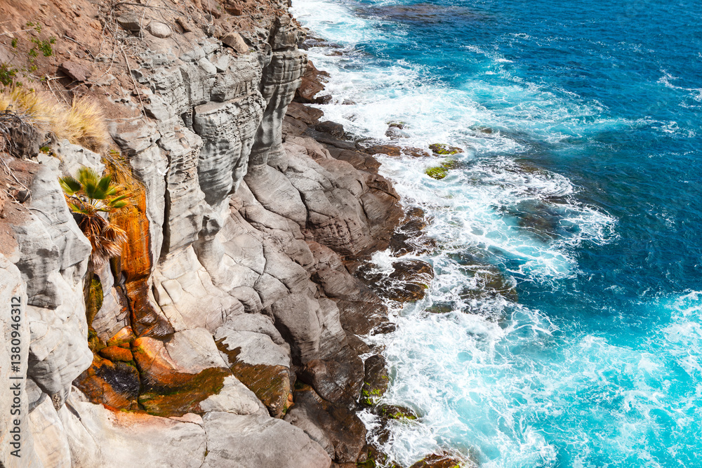 Dramatic coastal cliffs meet the turquoise ocean, waves crashing against the rugged rock face. Ocean waves surge against a textured cliff face, creating a dynamic coastal scene