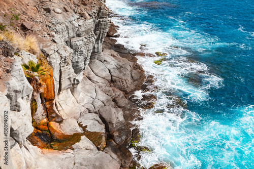Dramatic coastal cliffs meet the turquoise ocean, waves crashing against the rugged rock face. Ocean waves surge against a textured cliff face, creating a dynamic coastal scene