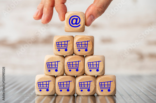 Eine Holzpyramide aus Würfel  ist bedruckt mit dem Symbol eines Einkaufswagen und eine Hand hält einen Holzwürfel in der Hand mit dem online-Symbol @