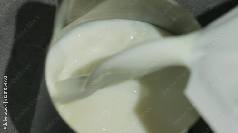 Milk pours into partially filled metal pitcher. Smooth stream of liquid cascades into container holding creamy substance slow motion
