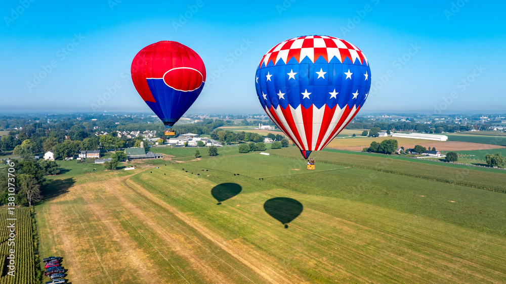 Obraz premium Two vibrant hot air balloons float gracefully above an expansive green field dotted with farms. The clear blue sky enhances the serene morning atmosphere.