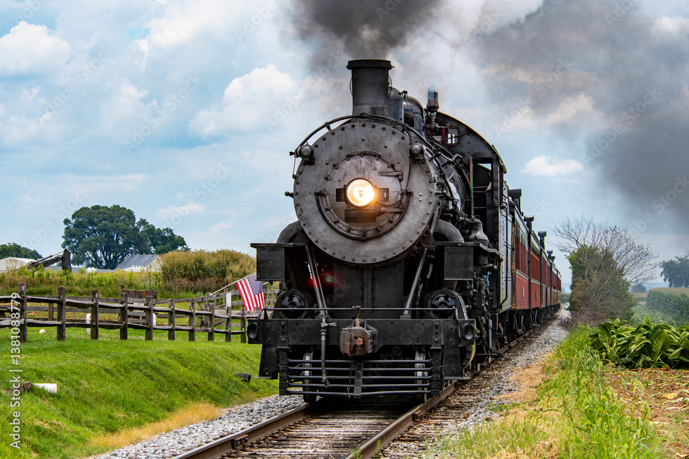 Fototapeta premium A historic steam locomotive travels along rural tracks, surrounded by lush greenery and waving American flags. The vibrant sky adds to the charm of this nostalgic journey.