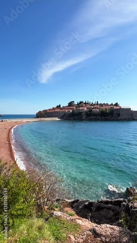 Scenic view of Sveti Stefan island in Montenegro, with clear turquoise waters, red rooftops, and a peaceful beach under a bright blue sky.