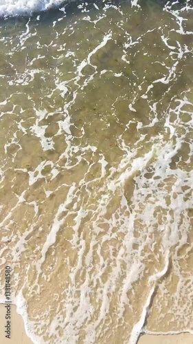  foamy sea waves gently washing over golden sand, creating dynamic textures and movement under sunlight in a peaceful coastal setting.