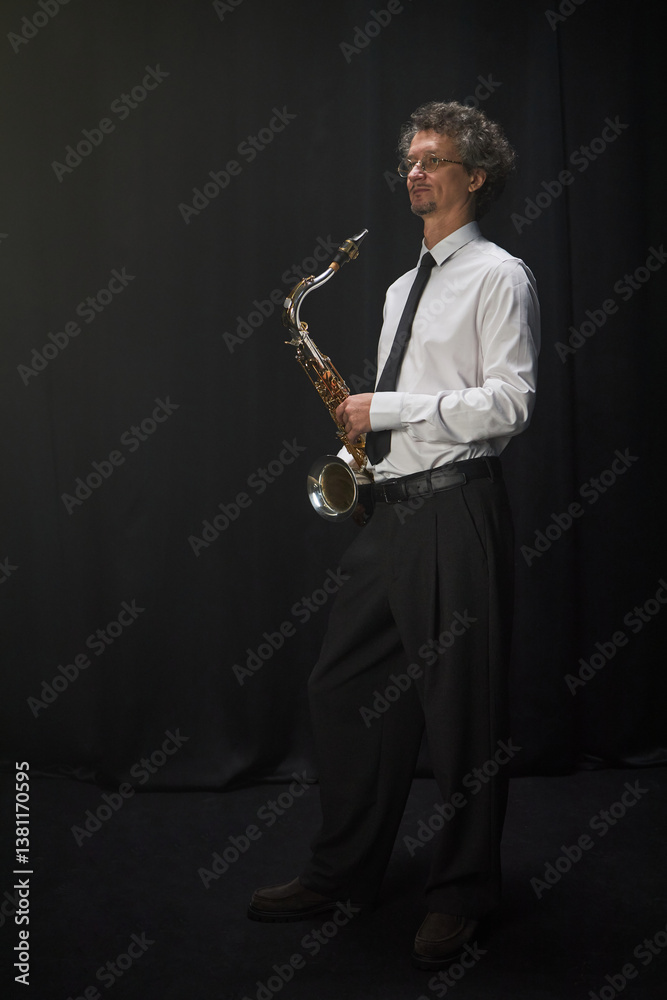 Naklejka premium Vertical studio shot of mature male musician dressed in formal concert attire posing with saxophone in hands standing against black curtain, copy space