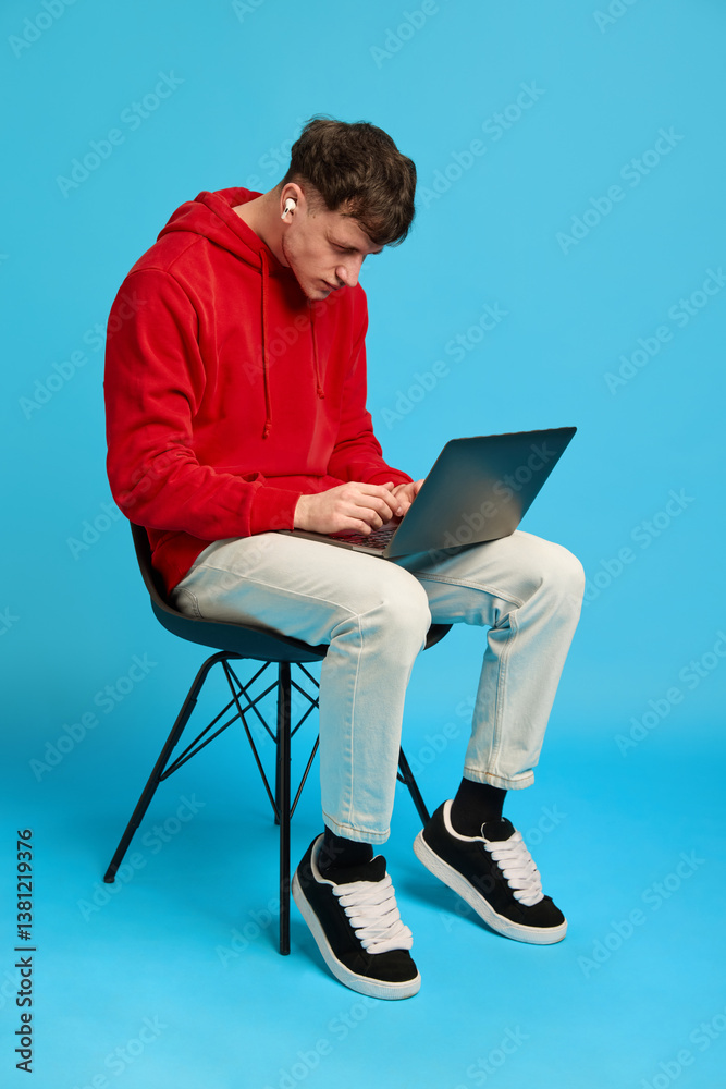 Focused young man with earbuds working on laptop while sitting on chair against blue studio background. Concept of remote work, digital business, freelance, self-expression, online life, style. Focused young man with earbuds working on laptop while sitting on chair against blue studio background. Concept of remote work, digital business, freelance, self-expression, online life, style.