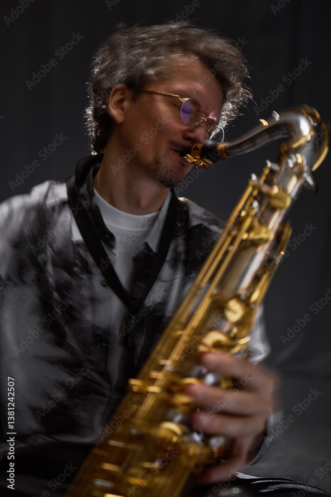 Fototapeta premium Vertical dynamic shot of professional male saxophonist in glasses playing jazz music with devotion against grey background in studio with backlight, slow shutter effect