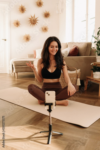 A fitness woman does sports at home using a mobile phone and a tripod while sitting on a exercise mat in a cozy interior. The influencer records an online workout. Healthy lifestyle. Selective focus