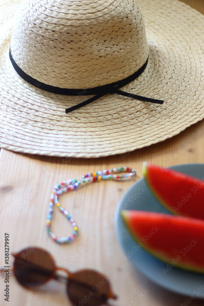 Plate with watermelon slices, glass of orange juice or cocktail, straw hat, colorful beaded necklace and sunglasses on the wooden table. Summer essentials concept. Selective focus.