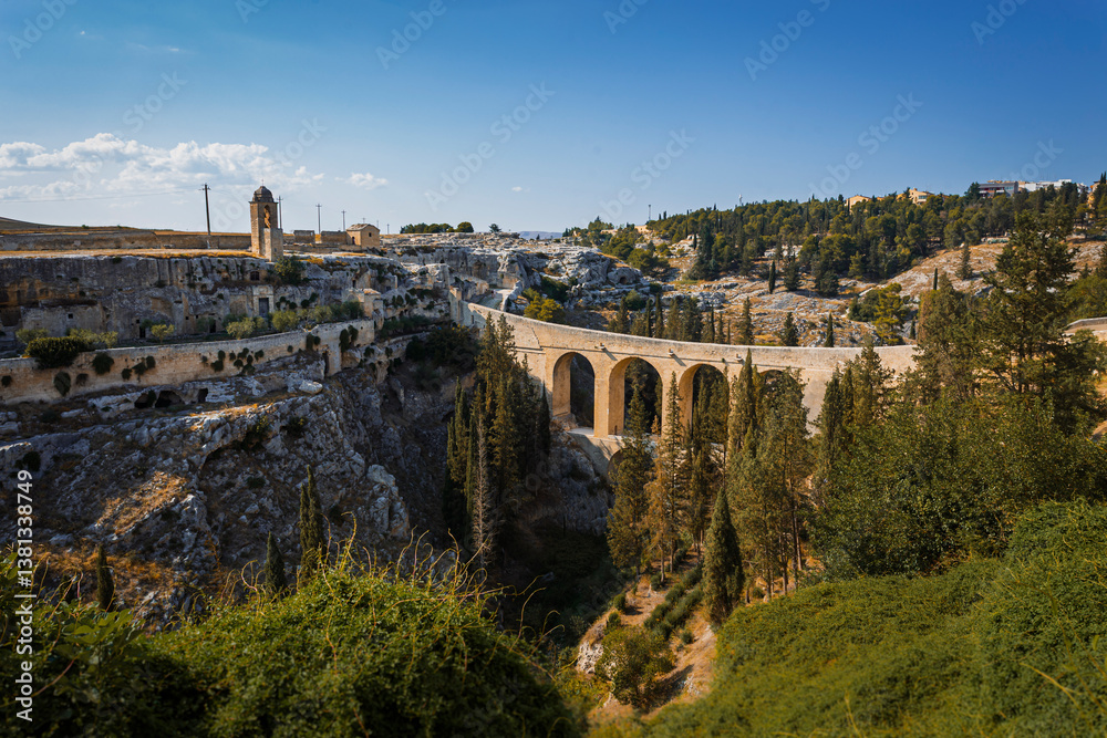 Fototapeta premium Scenic view of Gravina in Puglia, Italy, showcasing historic stone architecture, the iconic Roman bridge over the ravine, and the picturesque old town under a clear blue sky.